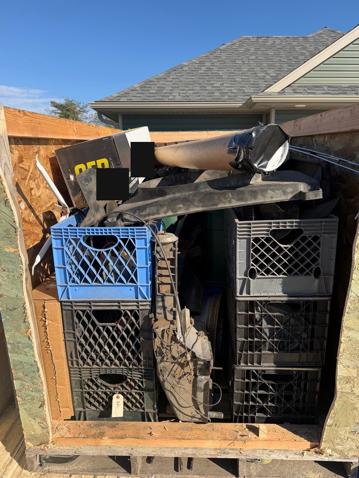 Open wooden crate holding six stacked plastic crates, boxes, a long wrapped item and several large loose parts. Shipping labels obscured.